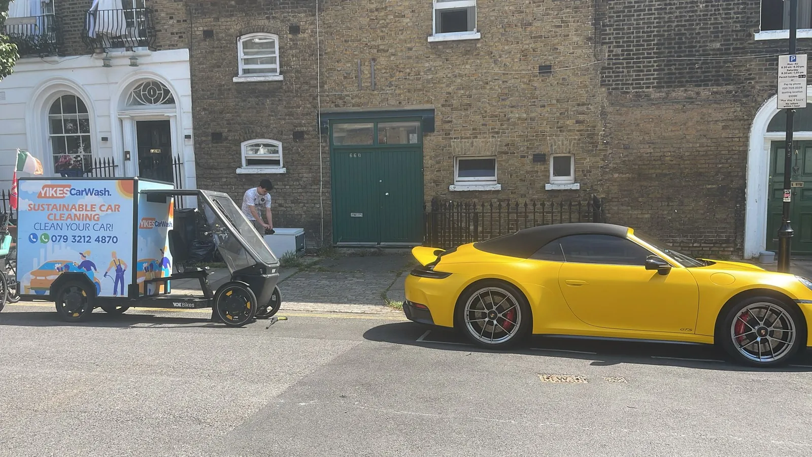 A yellow Porsche 911 GTS convertible at the kerb of a London street of Georgian houses, with a Yikes washer setting up beside it